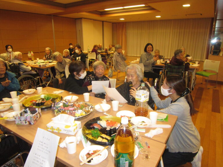 介護施設の食堂で複数の入居者が食事をしている様子。温かみのある空間で交流の時間を過ごしている。